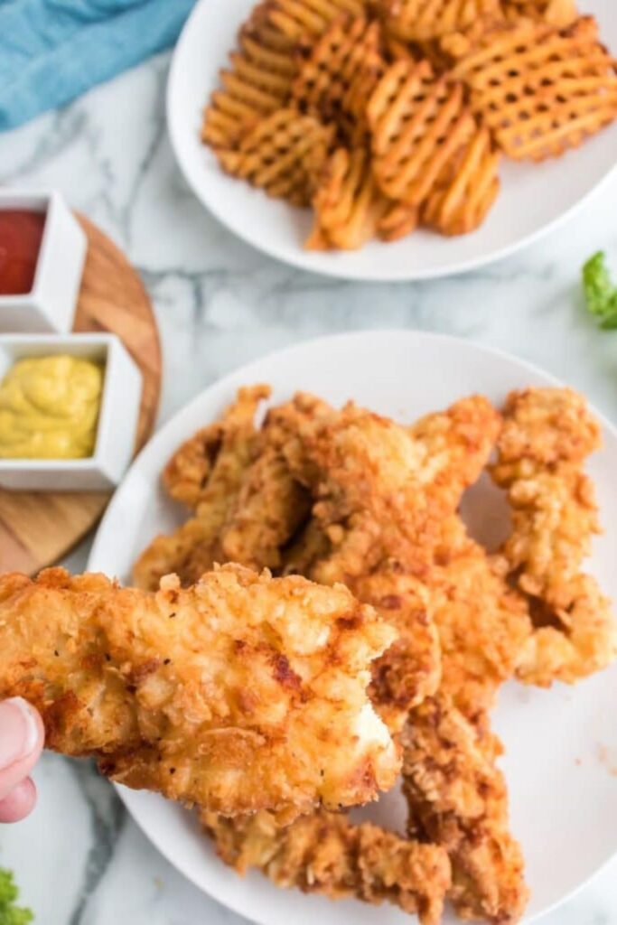 Homemade Chicken Strips on plate with one with a bite taken out of it being held in the hand