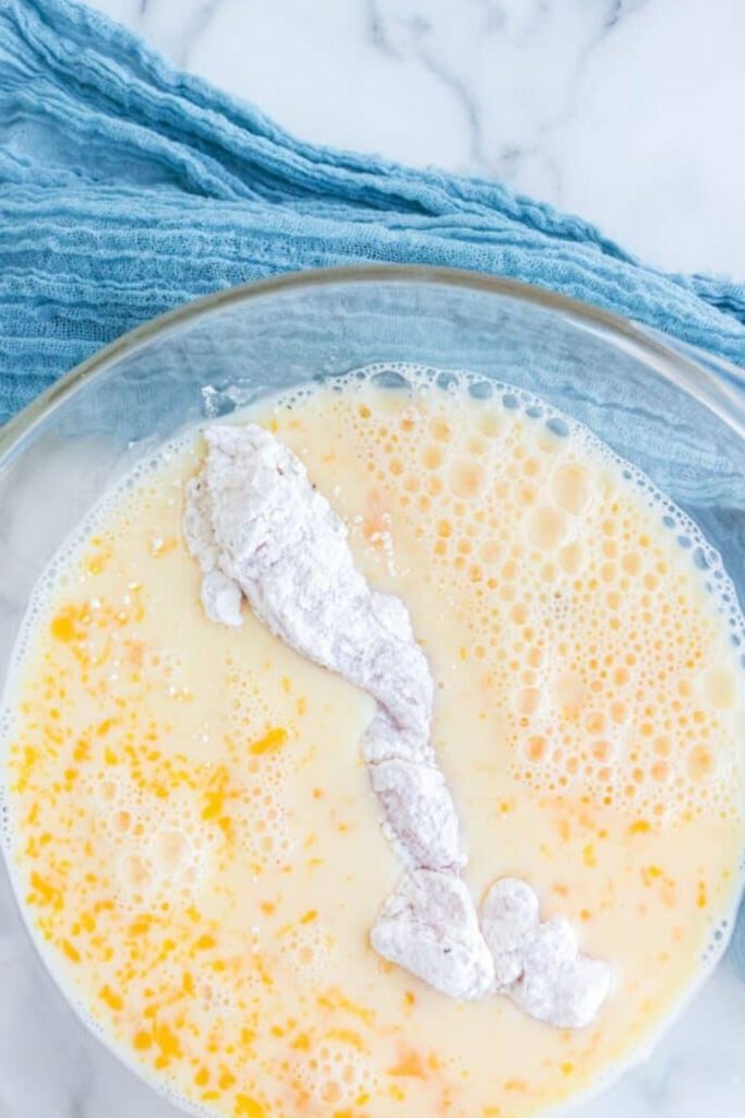 breaded chicken being dipped in an egg wash in a glass bowl sitting on table