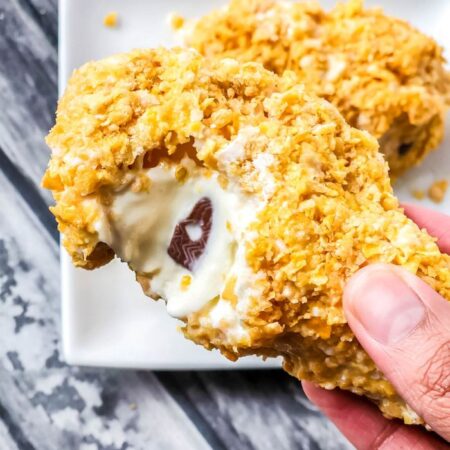 a chicken drumstick ice cream bar with bite taken out being held in a persons hand above wooden table