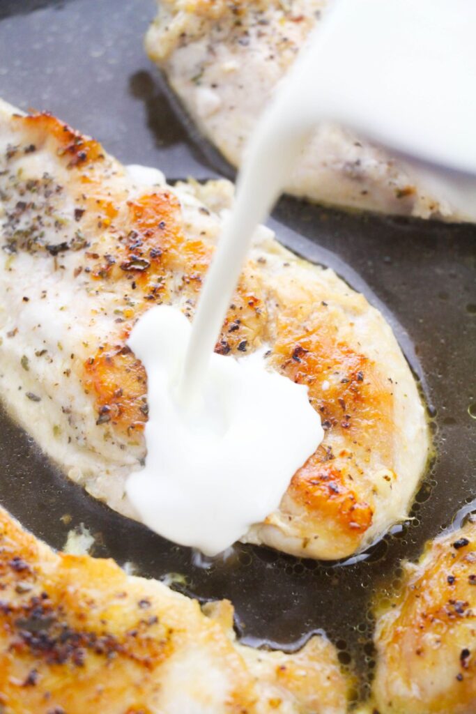 pouring heavy cream into a pan with seared chicken and chicken broth in it