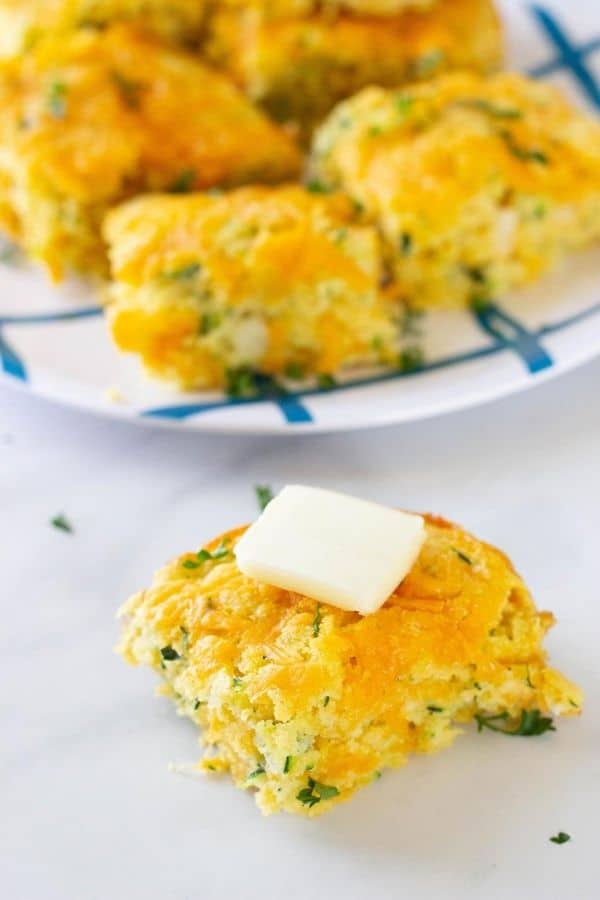 zucchini cornbread on table with a slab of butter on top. In background is a plate with cornbread stacked on it